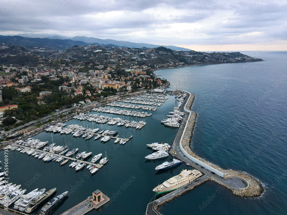 Fototapeta premium Aerial view of Sanremo, Italian city on the seashore in Liguria, north Italy. Drone flying along the port over beaches and boardwalk with palm trees and Birds Eye of yacht parking in San Remo, Italy.