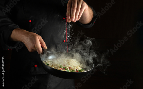 Fototapeta Naklejka Na Ścianę i Meble -  Professional chef adds salt to the spaghetti in a steaming hot pan. Recipe or menu for restaurant or hotel on black background