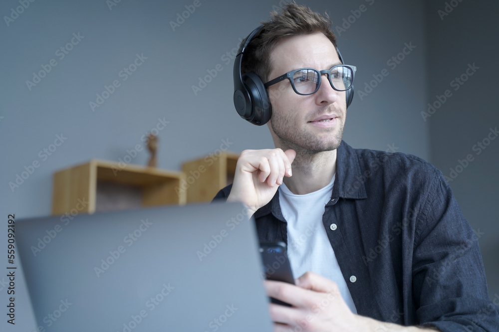 Thoughtful young businessman in headphones holds smartphone while sits at workplace at home