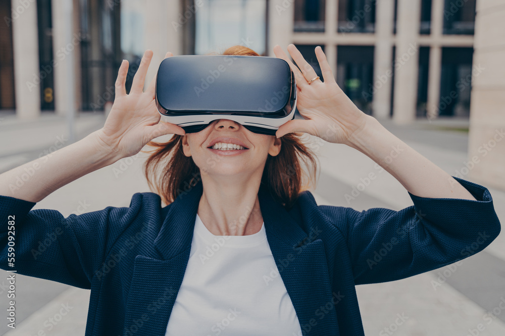 Young smiling redhead female entrepreneur testing portable VR goggles on city street Young smiling redhead female entrepreneur testing portable VR goggles on city street