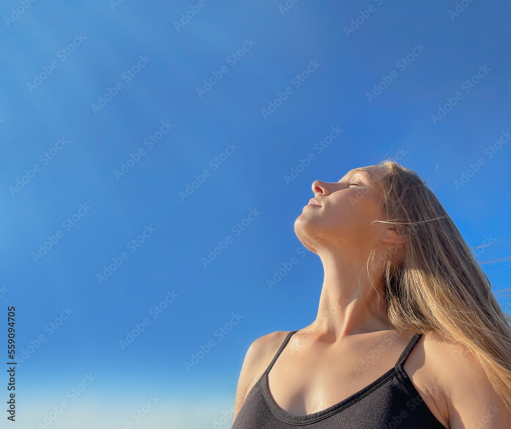 Portrait of a young relaxed girl, with long, light, loose hair, against a clear blue sunny sky, head turned to the rays of the sun, eyes closed. Sunbathing in the sun. Relaxes. Meditating.