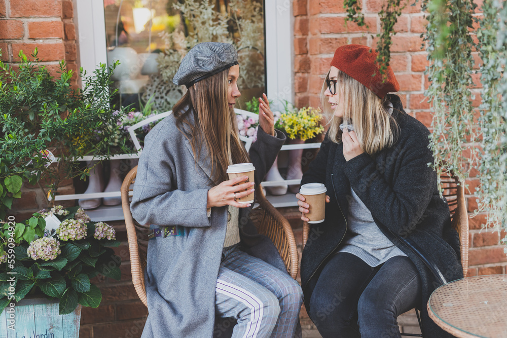 Fototapeta premium Young female best friends talking to each other discussing interesting themes during free time hipster girls joking and having fun together enjoying recreation with coffee to go on urban background
