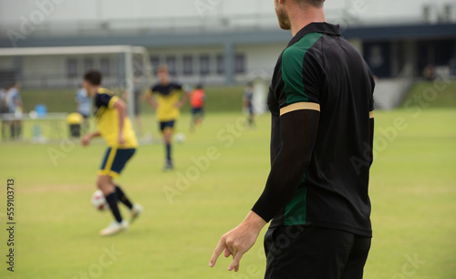 Male youth football coach standing and watching his team playing in a school tournament on a clear sky and sunny day. Sport, active lifestyle, happy activity and coaching concept.