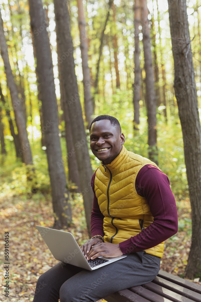 African american man sitting in city park on a bench with laptop studying online outdoors. Man freelancer learn working remotely in street e-learning vertical banner African american man sitting in city park on a bench with laptop studying online outdoors. Man freelancer learn working remotely in street e-learning vertical banner