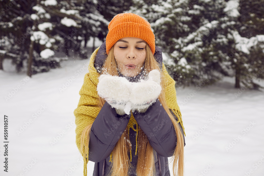 Fototapeta premium Winter mood. Portrait of beautiful young woman blowing snow from her palms during winter walk. Snowflakes fly from hands of girl in winter wear who walks in snowy forest and enjoys winter frosty day.