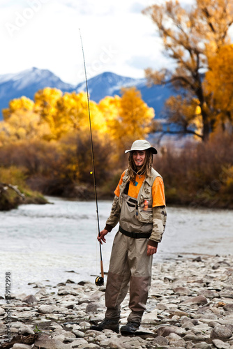 A athletic man fly fishing stands on the banks of a river with the fall colors and snowy mountains behind him in Montana. photo