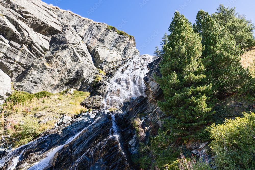 Multi-level alpine Lillaz waterfall (Cascate di Lillaz) washes granite karst rapids of rock covered with grass and evergreen trees under blue sky, Aosta valley, Italy