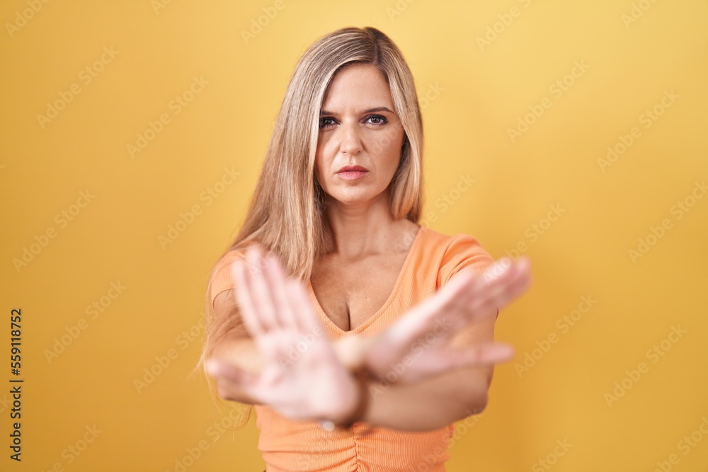 Young woman standing over yellow background rejection expression crossing arms and palms doing negative sign, angry face Young woman standing over yellow background rejection expression crossing arms and palms doing negative sign, angry face