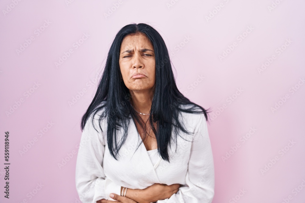 Mature hispanic woman standing over pink background with hand on stomach because indigestion, painful illness feeling unwell. ache concept. Mature hispanic woman standing over pink background with hand on stomach because indigestion, painful illness feeling unwell. ache concept.