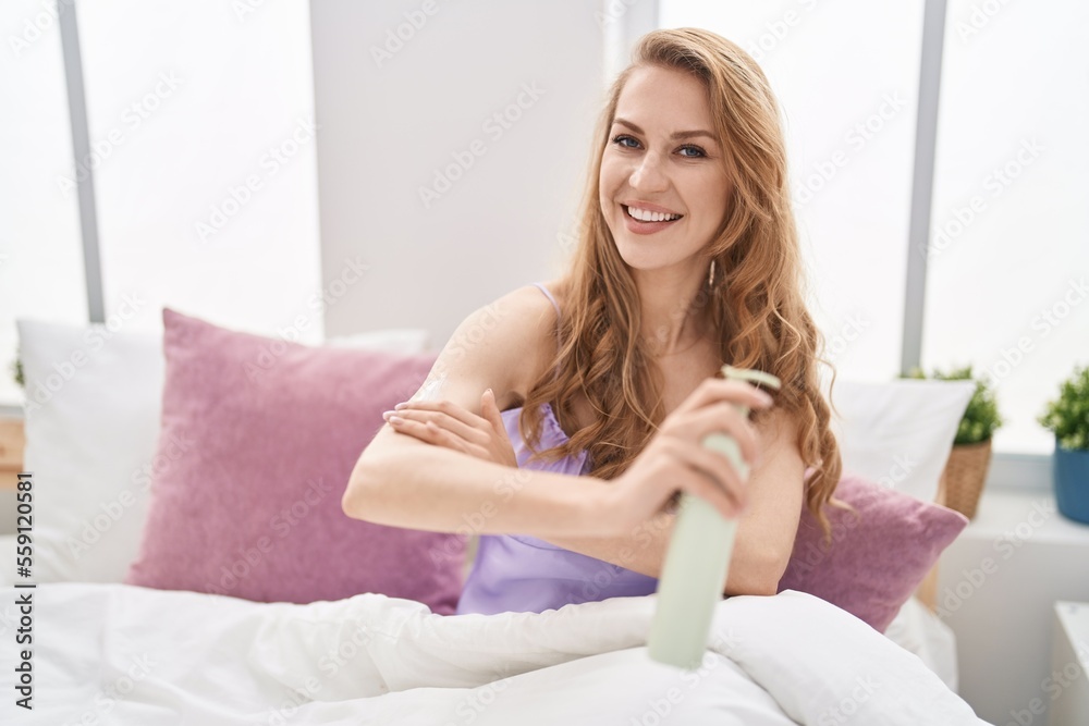 Young blonde woman applying skin lotion sitting on bed at bedroom Young blonde woman applying skin lotion sitting on bed at bedroom