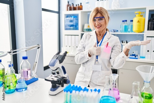 Middle age blonde woman working at scientist laboratory showing palm hand and doing ok gesture with thumbs up  smiling happy and cheerful