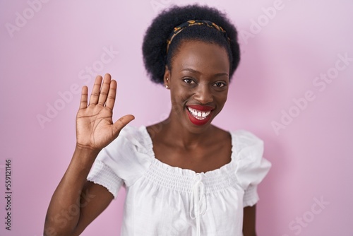 African woman with curly hair standing over pink background waiving saying hello happy and smiling, friendly welcome gesture