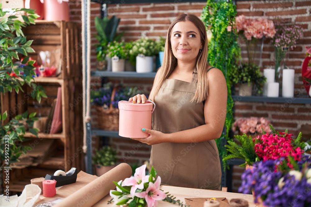 Young blonde woman working at florist shop smiling looking to the side and staring away thinking.