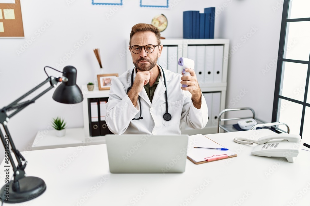 Middle age doctor man holding thermometer at the clinic serious face thinking about question with hand on chin, thoughtful about confusing idea Middle age doctor man holding thermometer at the clinic serious face thinking about question with hand on chin, thoughtful about confusing idea