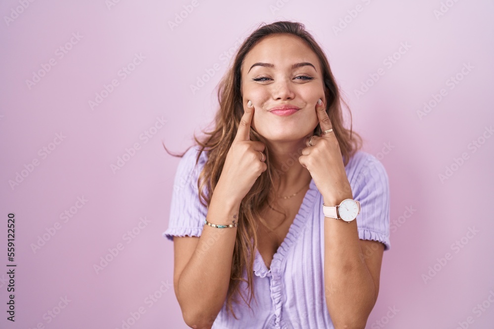 Young hispanic woman standing over pink background smiling with open mouth, fingers pointing and forcing cheerful smile Young hispanic woman standing over pink background smiling with open mouth, fingers pointing and forcing cheerful smile