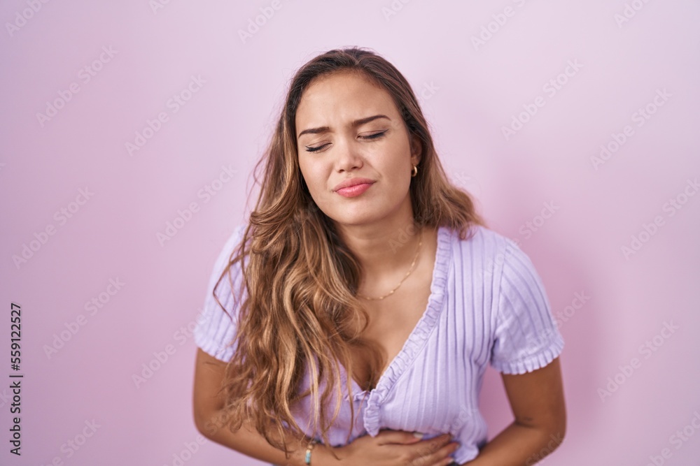 Young hispanic woman standing over pink background with hand on stomach because indigestion, painful illness feeling unwell. ache concept. Young hispanic woman standing over pink background with hand on stomach because indigestion, painful illness feeling unwell. ache concept.