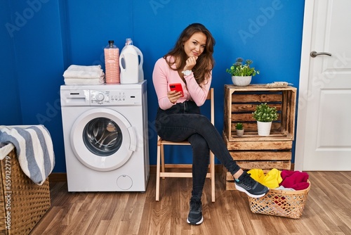 Young hispanic woman sitting waiting for laundry using smartphone looking confident at the camera smiling with crossed arms and hand raised on chin. thinking positive.