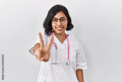 Young hispanic doctor woman wearing stethoscope over isolated background smiling looking to the camera showing fingers doing victory sign. number two.