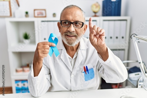 Mature doctor man holding blue ribbon at the clinic surprised with an idea or question pointing finger with happy face  number one