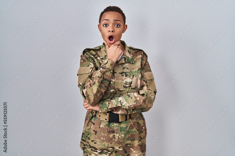Beautiful african american woman wearing camouflage army uniform looking fascinated with disbelief, surprise and amazed expression with hands on chin