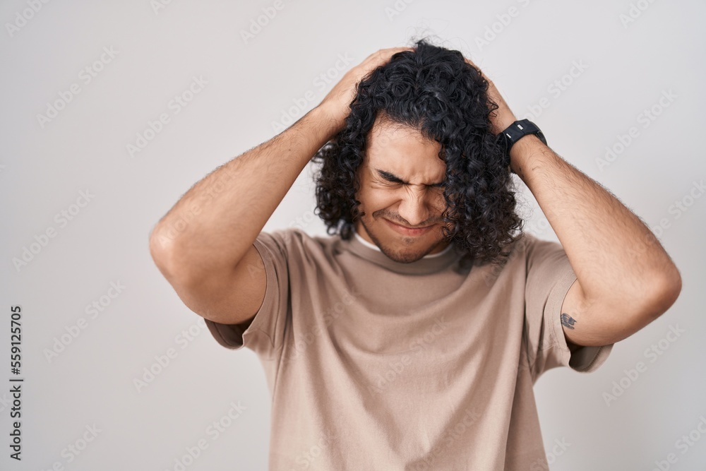 Hispanic man with curly hair standing over white background suffering from headache desperate and stressed because pain and migraine. hands on head.