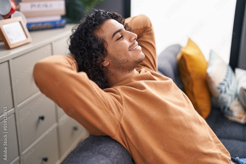 Fototapeta premium Young latin man relaxed with hands on head sitting on sofa at home