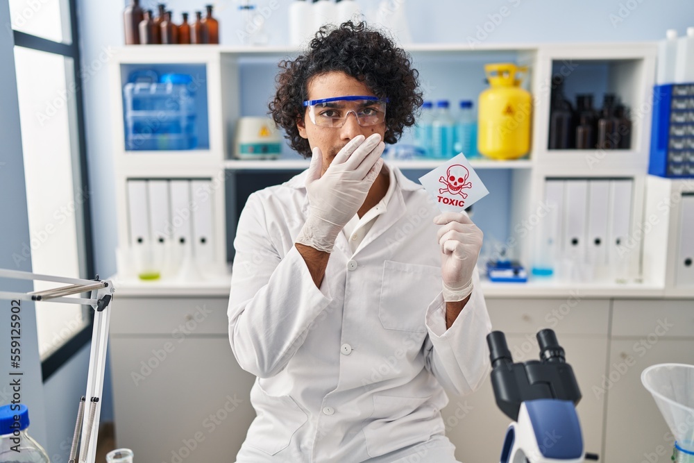 Hispanic man with curly hair working at scientist laboratory holding toxic banner covering mouth with hand, shocked and afraid for mistake. surprised expression Hispanic man with curly hair working at scientist laboratory holding toxic banner covering mouth with hand, shocked and afraid for mistake. surprised expression