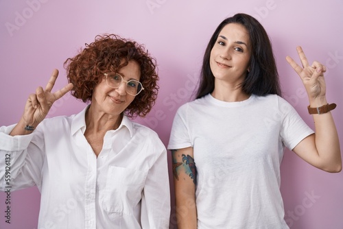 Hispanic mother and daughter wearing casual white t shirt over pink background smiling looking to the camera showing fingers doing victory sign. number two.