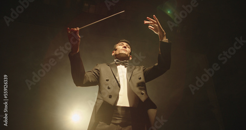Asian symphony orchestra conductor wearing suit is directing musicians with movement of baton, isolated on black smokey background photo