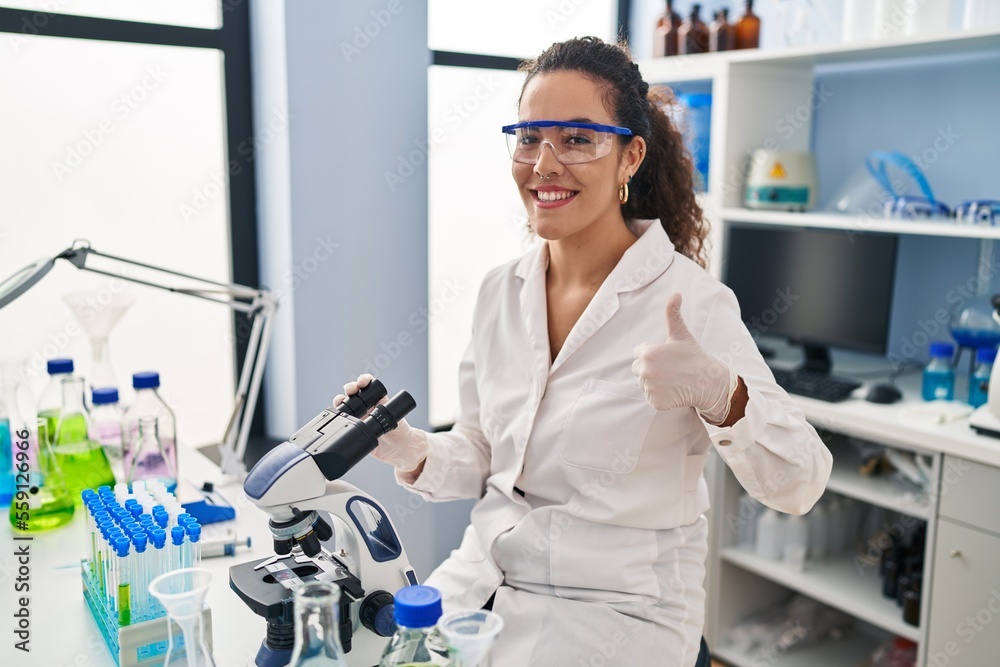 Young hispanic woman working at scientist laboratory smiling happy and positive, thumb up doing excellent and approval sign