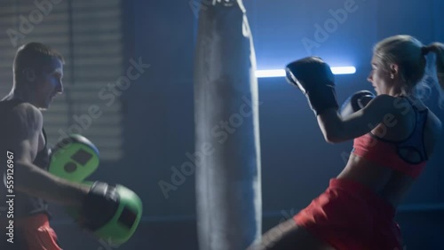 Kickboxing training. Woman in boxing gloves is boxing with her coach in dark gym with blue light. Training sparring of female kickboxer and male trainer. Female fighter practices punches and kicks.