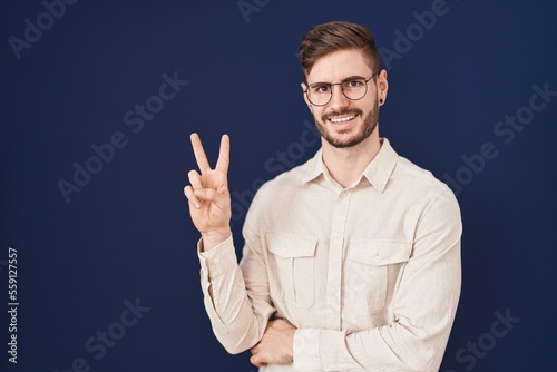 Hispanic man with beard standing over blue background smiling with happy face winking at the camera doing victory sign. number two.