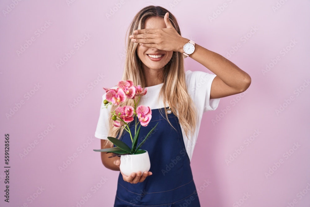 Young blonde woman wearing gardener apron holding plant smiling and laughing with hand on face covering eyes for surprise. blind concept.