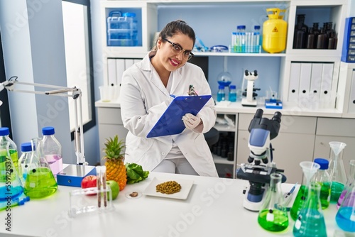 Young beautiful plus size woman scientist smiling confident writing report working at pharmacy