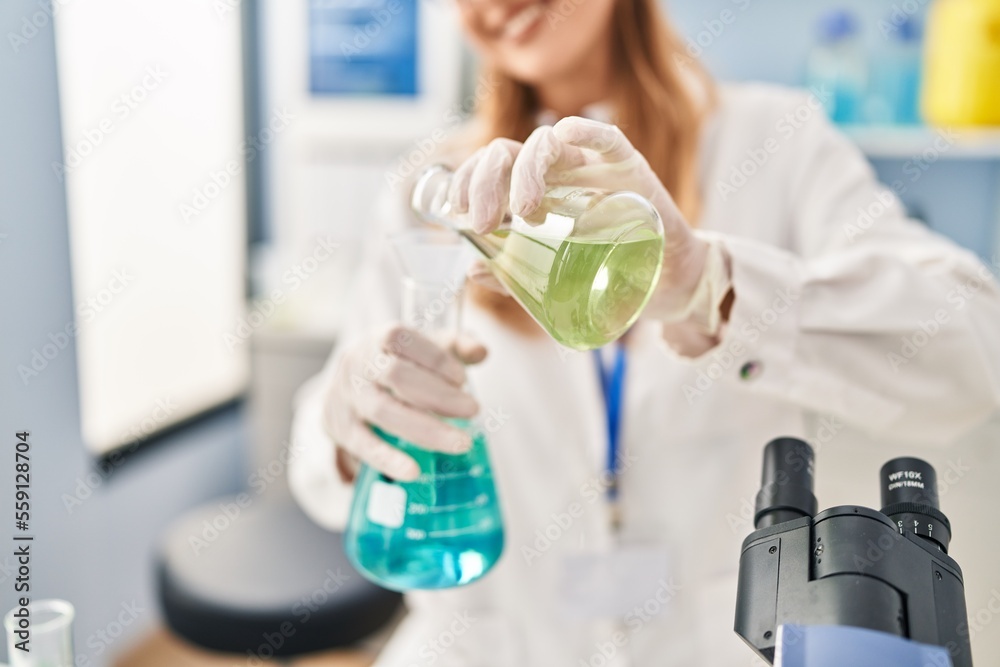 Fototapeta premium Young blonde woman wearing scientist uniform pouring liquid on test tube at laboratory