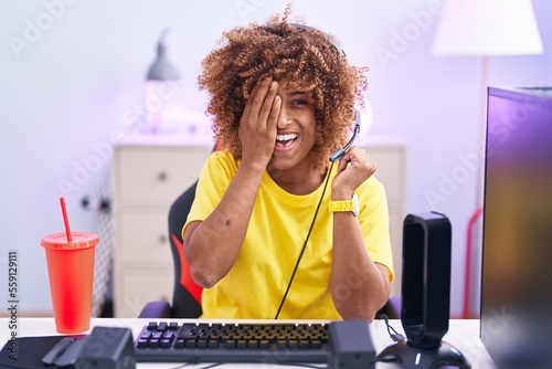 Young hispanic woman with curly hair playing video games wearing headphones covering one eye with hand  confident smile on face and surprise emotion.