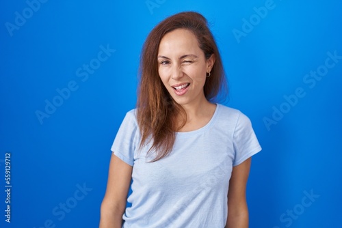 Brunette woman standing over blue background winking looking at the camera with sexy expression  cheerful and happy face.