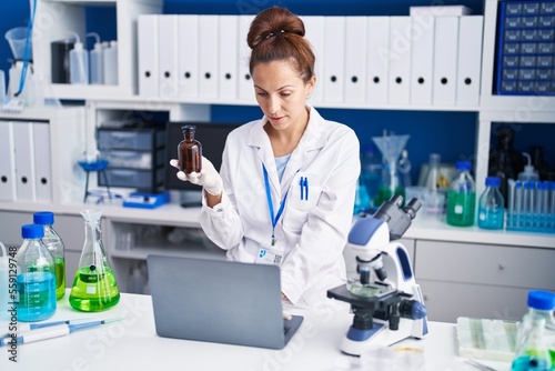 Young woman scientist using laptop holding bottle at laboratory