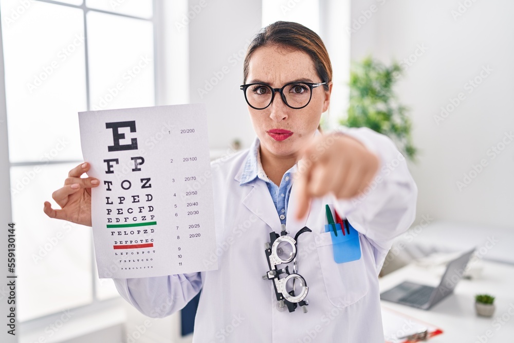 Young hispanic optician woman holding medical exam pointing with finger to the camera and to you, confident gesture looking serious Young hispanic optician woman holding medical exam pointing with finger to the camera and to you, confident gesture looking serious