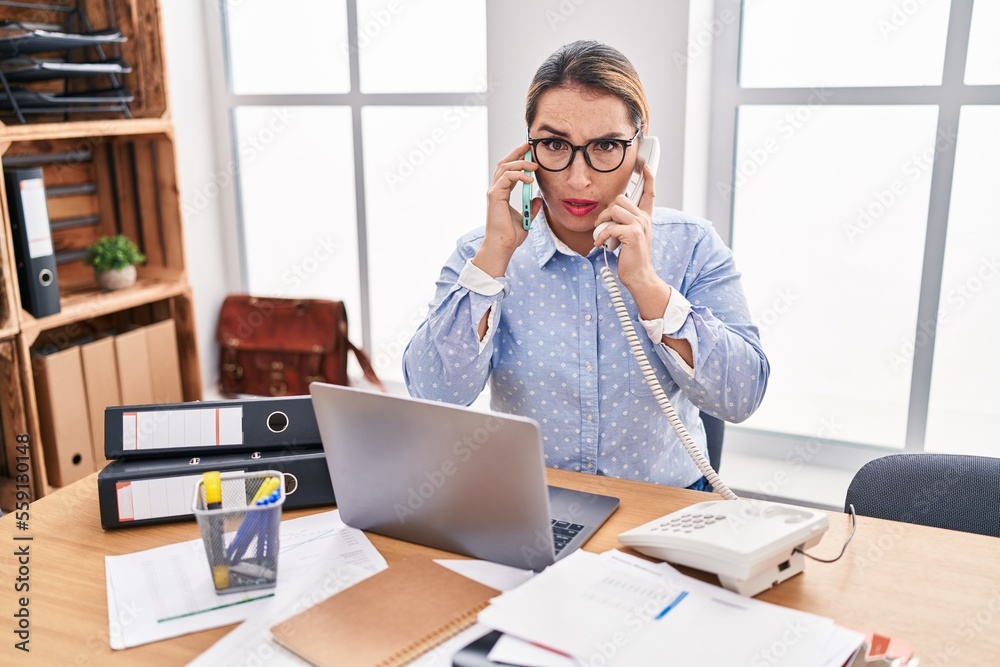 Young hispanic business woman working at the office talking on two phones skeptic and nervous, frowning upset because of problem. negative person. Young hispanic business woman working at the office talking on two phones skeptic and nervous, frowning upset because of problem. negative person.