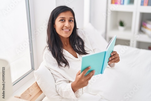 Young beautiful hispanic woman reading book sitting on bed at bedroom