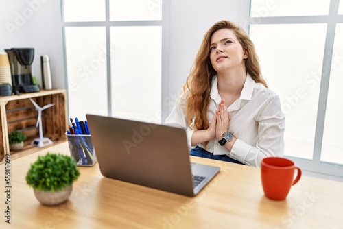 Young caucasian woman working at the office using computer laptop begging and praying with hands together with hope expression on face very emotional and worried. begging. © Krakenimages.com