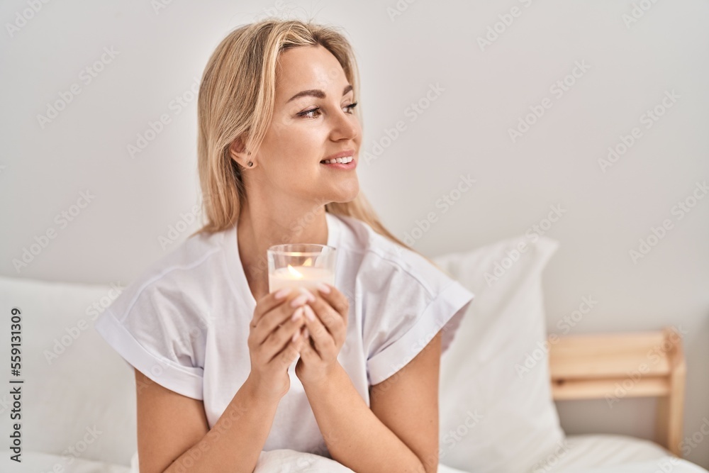 Young blonde woman drinking glass of water sitting on bed at bedroom