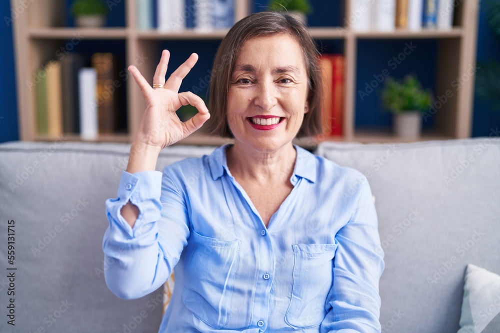 Middle age hispanic woman sitting on the sofa at home smiling positive doing ok sign with hand and fingers. successful expression. Middle age hispanic woman sitting on the sofa at home smiling positive doing ok sign with hand and fingers. successful expression.