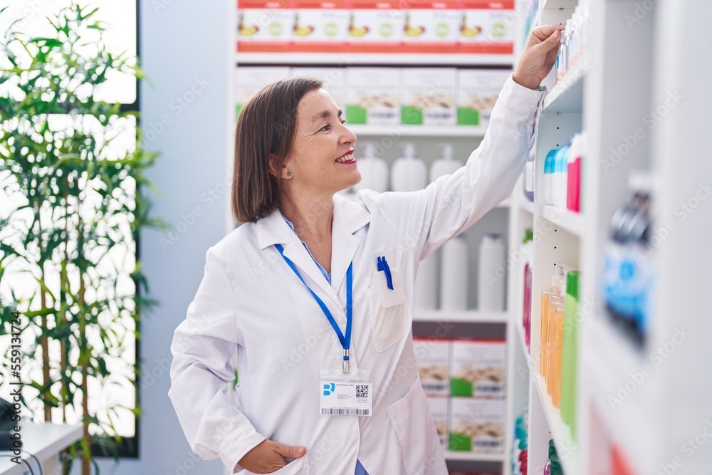 Middle age woman pharmacist smiling confident holding product on shelving at pharmacy