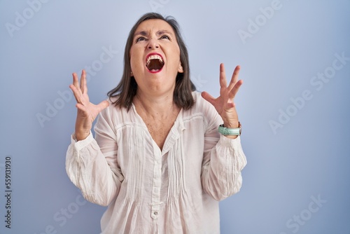 Middle age hispanic woman standing over blue background crazy and mad shouting and yelling with aggressive expression and arms raised. frustration concept.