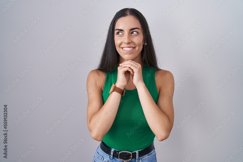 Young woman standing over isolated background laughing nervous and excited with hands on chin looking to the side Young woman standing over isolated background laughing nervous and excited with hands on chin looking to the side