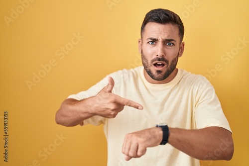 Handsome hispanic man standing over yellow background in hurry pointing to watch time, impatience, upset and angry for deadline delay photo
