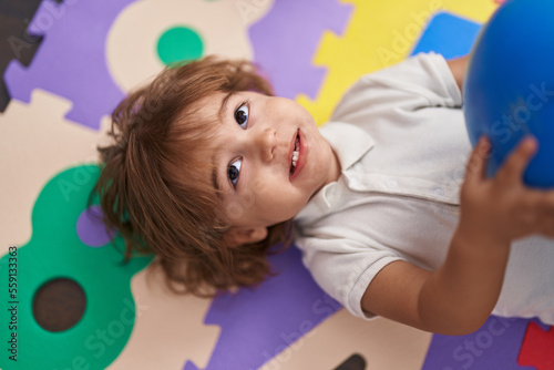 Adorable hispanic toddler holding ball lying on floor at kindergarten