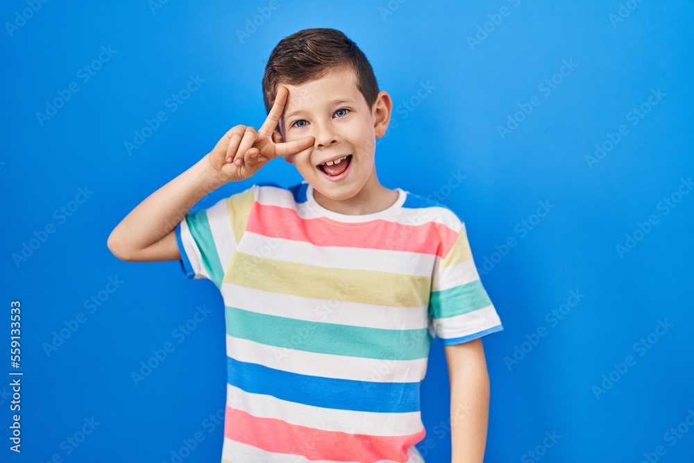 Young caucasian kid standing over blue background doing peace symbol with fingers over face, smiling cheerful showing victory Young caucasian kid standing over blue background doing peace symbol with fingers over face, smiling cheerful showing victory